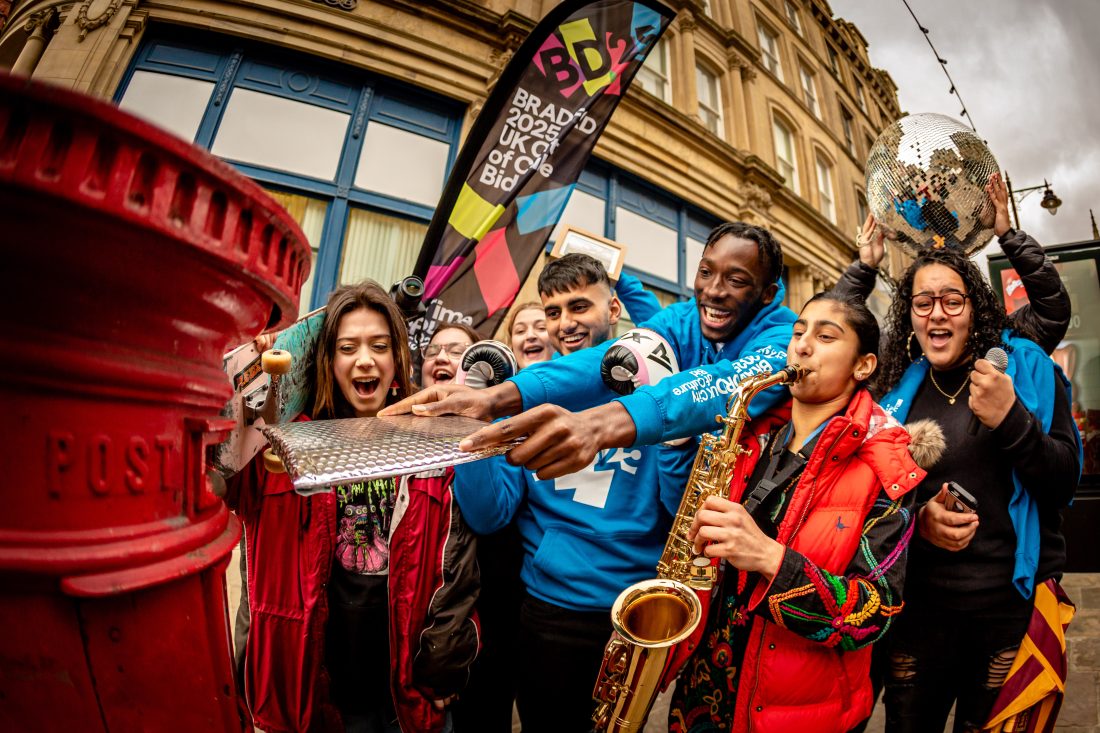 A group of young people gather on a city street around a red post box, while one person plays a saxophone and others watch and smile as they post an item in silver packaging