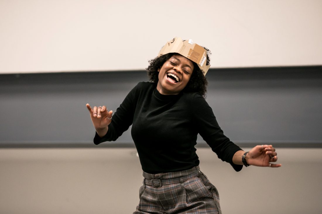Photo of a Black actor rehearsing in character, wearing a cardboard crown and smiling with her eyes closed.