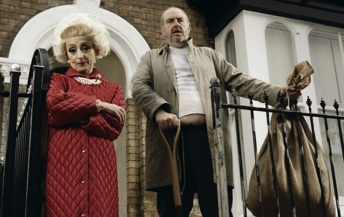 Photo of a man and woman in character in front of a house. The woman wears a long, red coat and the man holds a spade and a large sack.