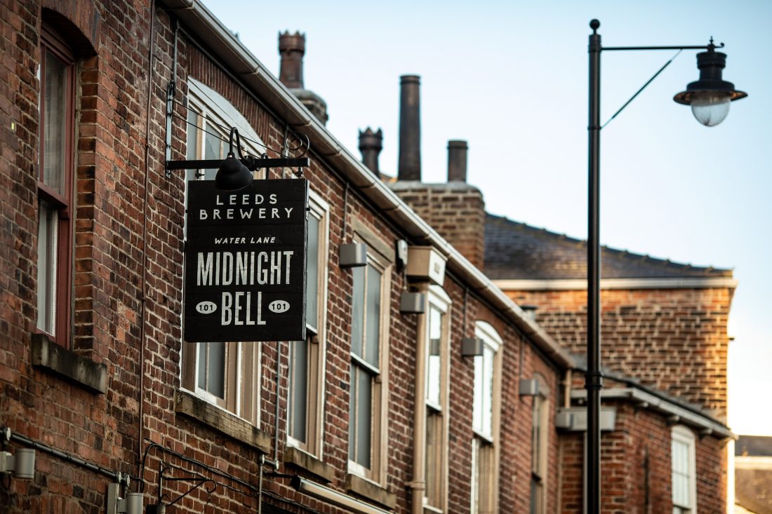 A black hanging sign reading “Leeds Brewery – Midnight Bell” is mounted on the exterior of a brick building along a narrow street.