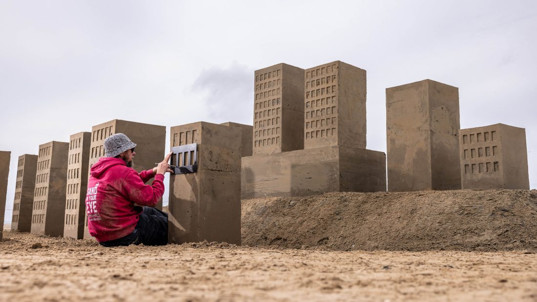 Photo of a sculptor in a red hoodie creating large, uniform and very precise tower blocks made from sand.