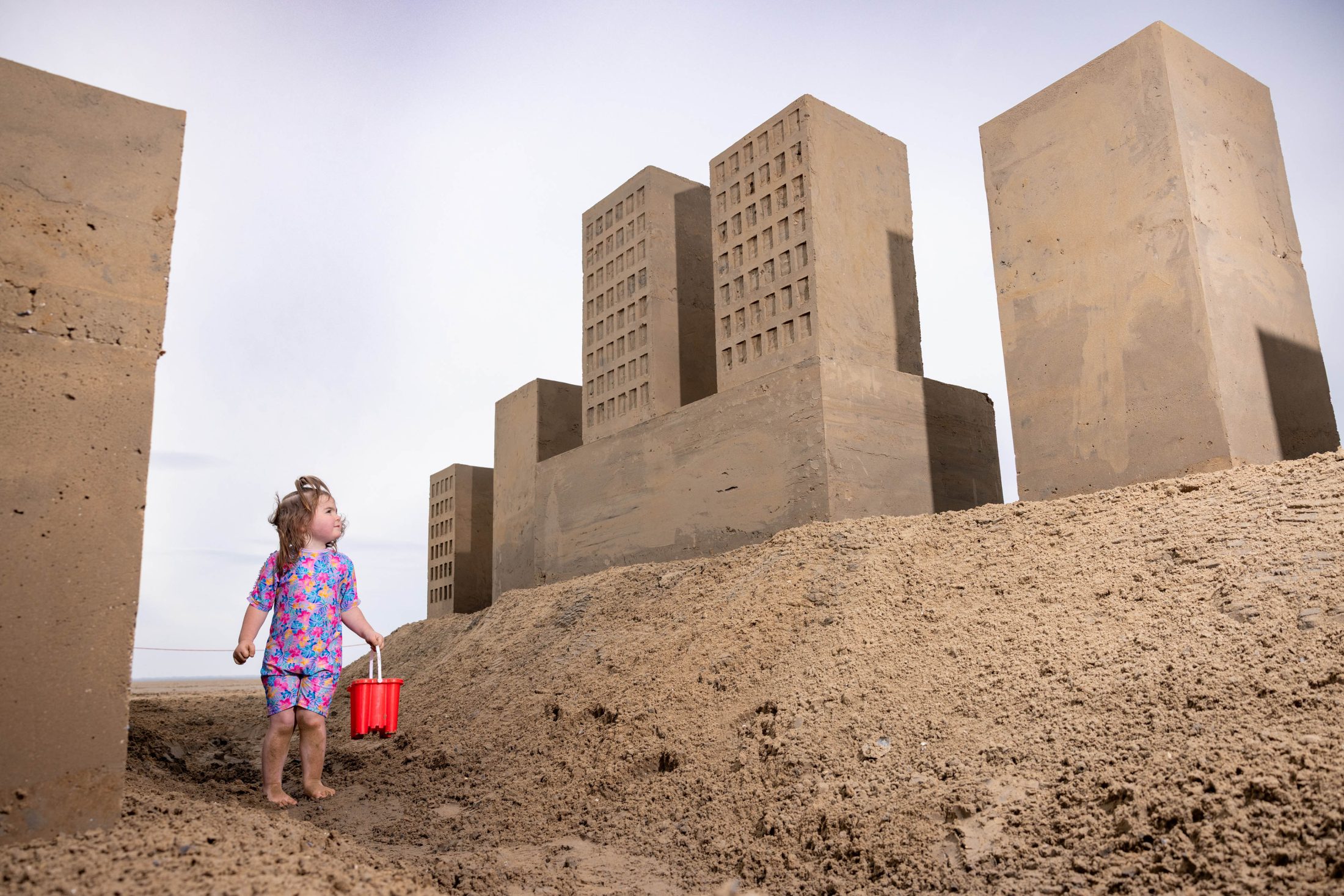 Photo of a toddler holding a red bucket on a beach, surrounded by large, uniform and very precise tower blocks made from sand.