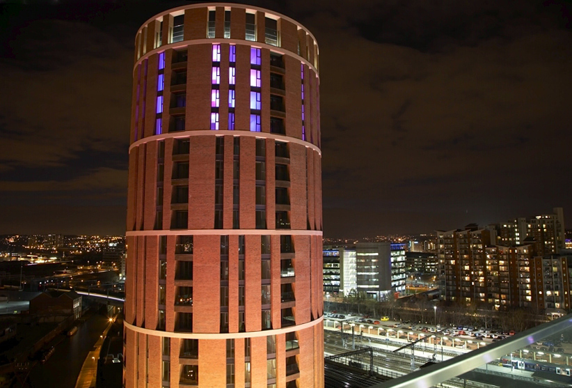 Aerial photo of Candle House – a tall, cylindrical, red brick building with balconies – lit up at night.