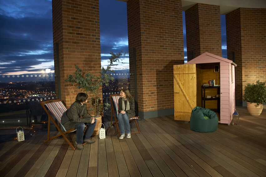 Photo of two people sitting on deck chairs inside. Views from floor-to-ceiling windows show they’re high up and it’s twilight. There are potted plants and lanterns on the wooden floor, and a light-pink shed with books inside stands open to the right