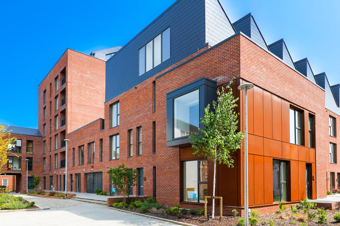 A contemporary red-brick residential building with corten steel cladding and large windows stands along a pedestrian walkway with trees and planted areas.