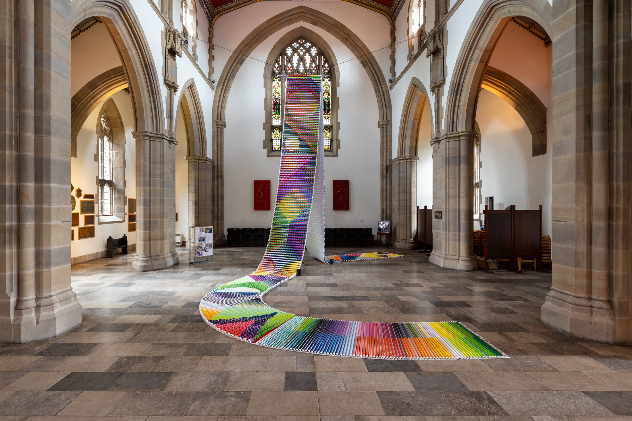 Photo of a brightly coloured, path-like sculpture hanging from a line in a church and trailing to the floor.