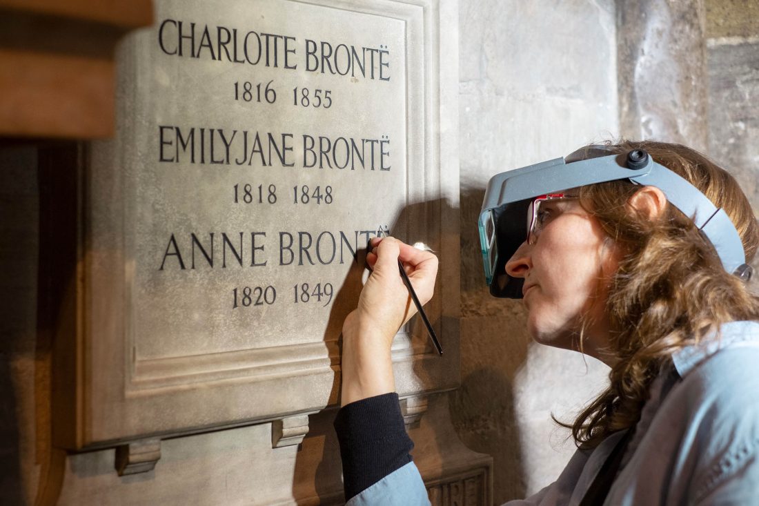 Photo of a stone plaque with the Brontë sisters’ names and dates on. A painter is adding dots above the final ‘E’.