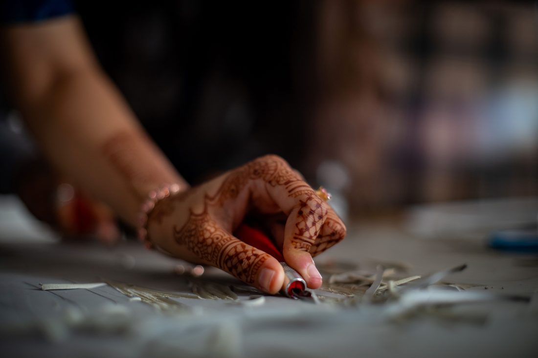Close-up photo of a hand, decorated with henna, chiselling chips off a flat piece of wood.