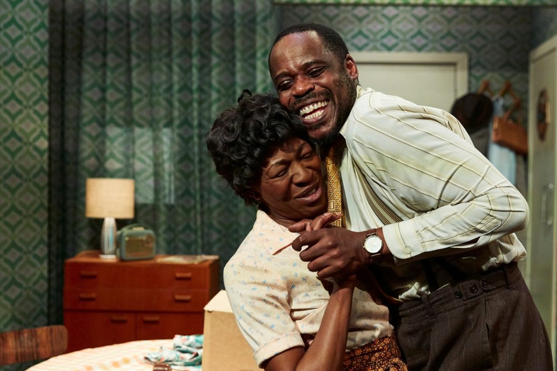 Photo of two Black actors in character – a man and a woman embracing happily with the set of a home behind them