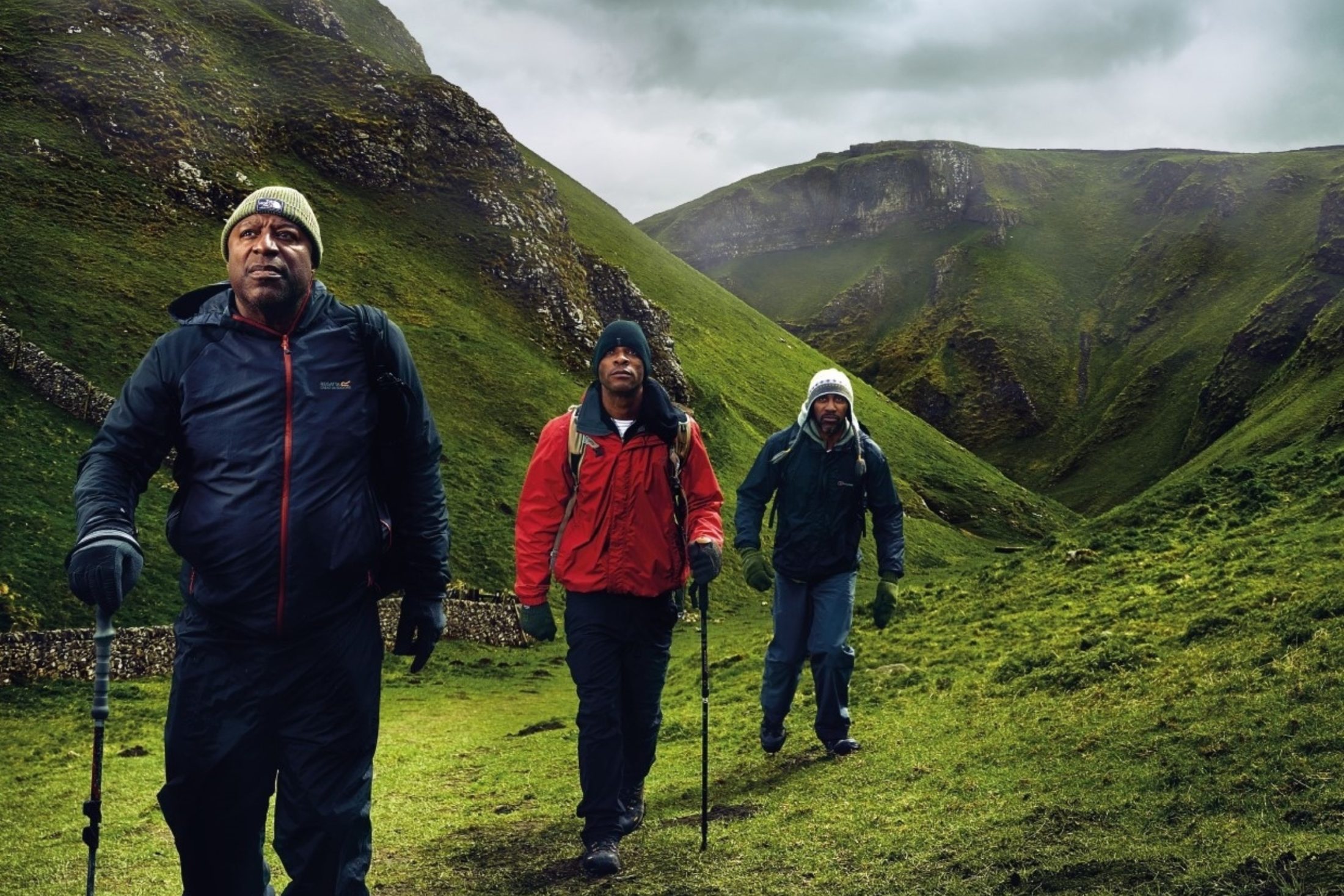 Photo of three Black actors, in character, hiking through green mountains with stormy clouds above them.