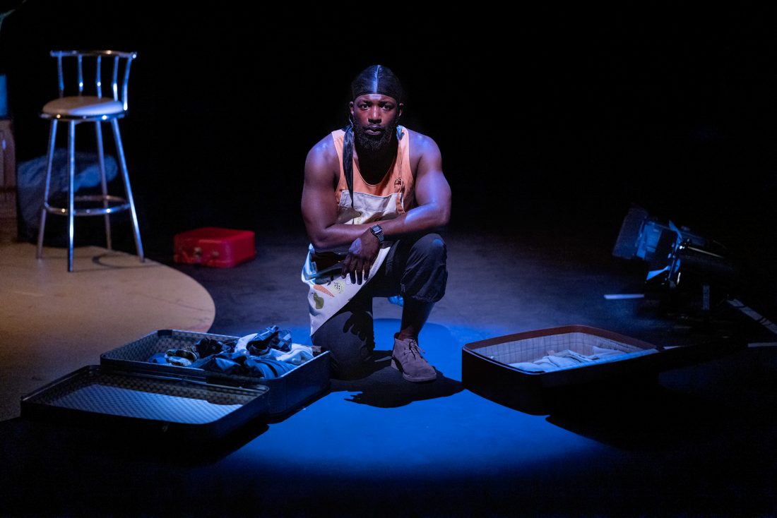 Photo of an actor in character, kneeling in a blue spotlight on a stage, surrounded by open suitcases.