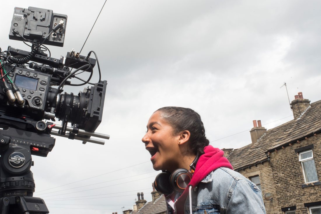 Photo of a young woman from the shoulders up, shouting gleefully into a professional filming camera. Old terraced houses are just visible behind her.