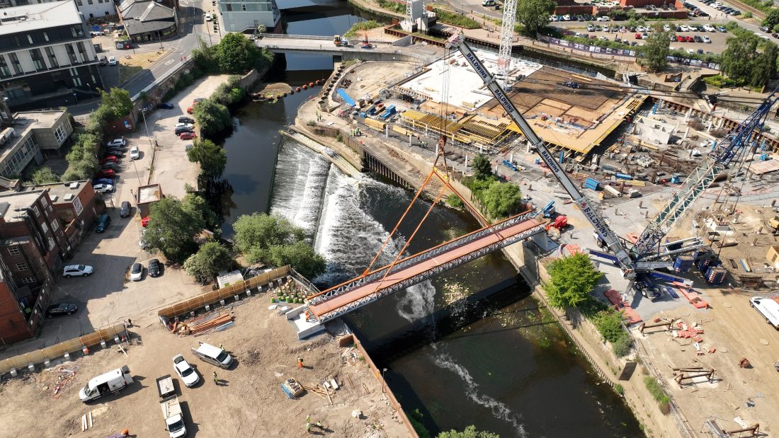 Aerial photo of the Forge Island bridge being lifted into place by a crane over a river. Behind it, building work is being carried out.