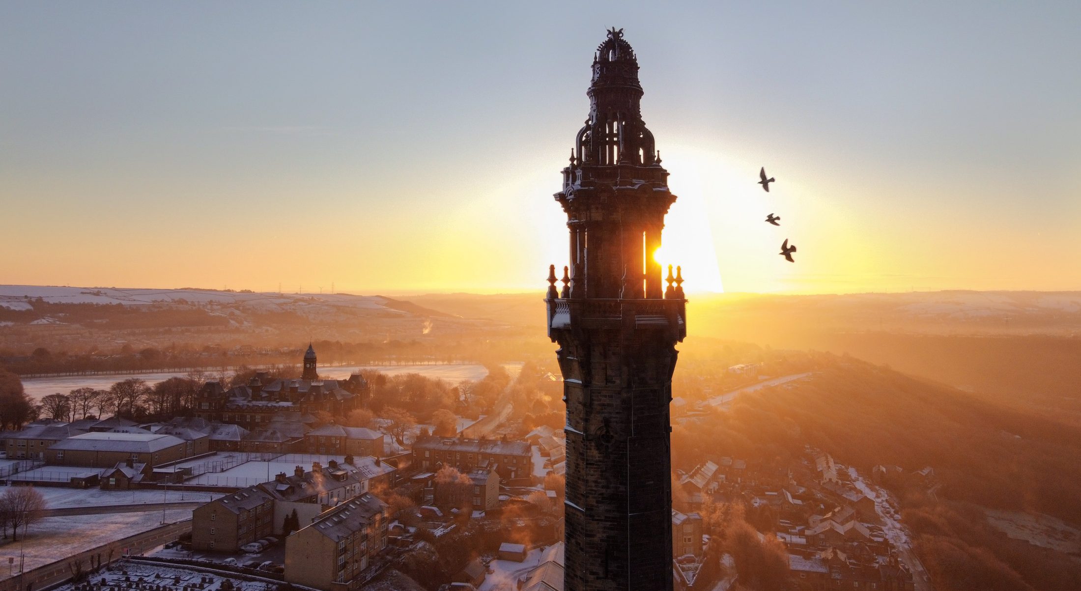 Aerial photo of the ornate Wainhouse Tower and three swooping birds, silhouetted against a wintry sunrise.