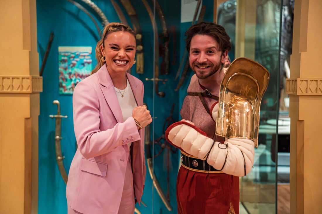 Photo of a woman in a pink blazer and a man dressed as a gladiator smiling at the camera with biceps curled.