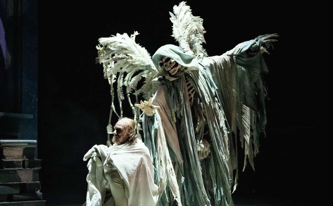 Photo of a ballet dancer in character as Scrooge on stage, with another dancer dressed as an angel of death creeping up behind him.