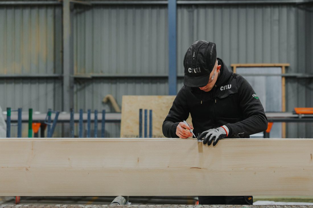 Photo of a Citu worker in a black cap and sweater, carefully measuring a length of timber in a warehouse.