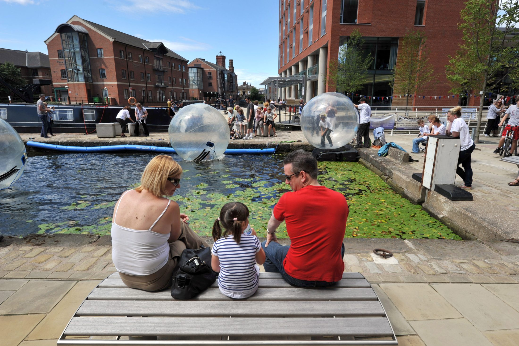 Photo taken from behind a couple with a child between them, sitting on a wooden bench facing the docks on a sunny day. In the background are red-brick warehouses and people zorbing in inflatable balls on the docks.