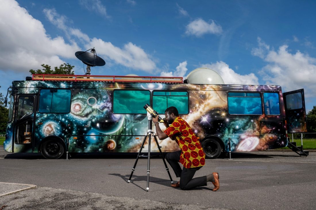Photo of a man looking through a telescope on a blue-sky day. In the background is a bus decorated with a galaxy print.