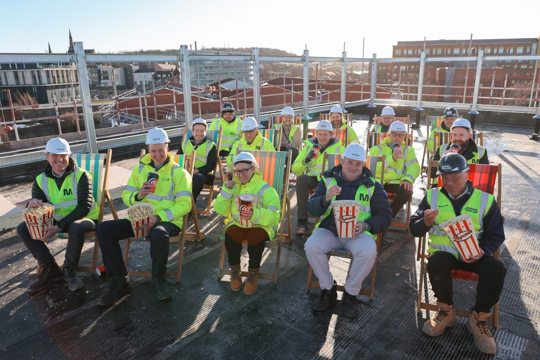 Photo of a group of people in fluorescent jackets and hard hats, sitting on striped deck chairs outside, holding tubs of popcorn and smiling at the camera.