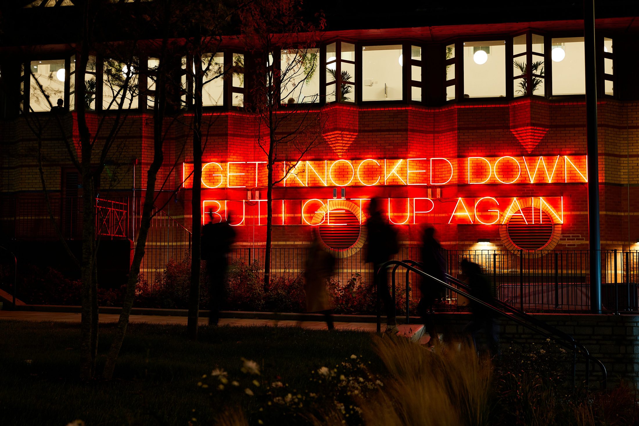 Photo of the side of Leeds Playhouse at night, with neon red lights on the wall, reading ‘I get knocked down but I get up again’.