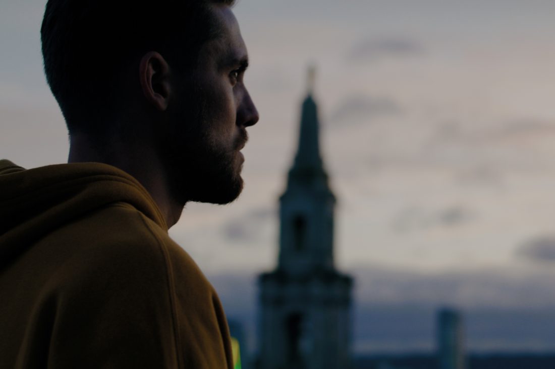 Photo of a man in a yellow hoodie, looking out across the Leeds skyline at dusk.