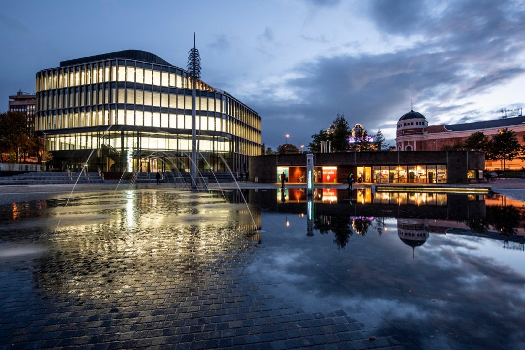 Photo of One City Park lit up at twilight, reflected in the water of the Mirror Pool in Bradford.