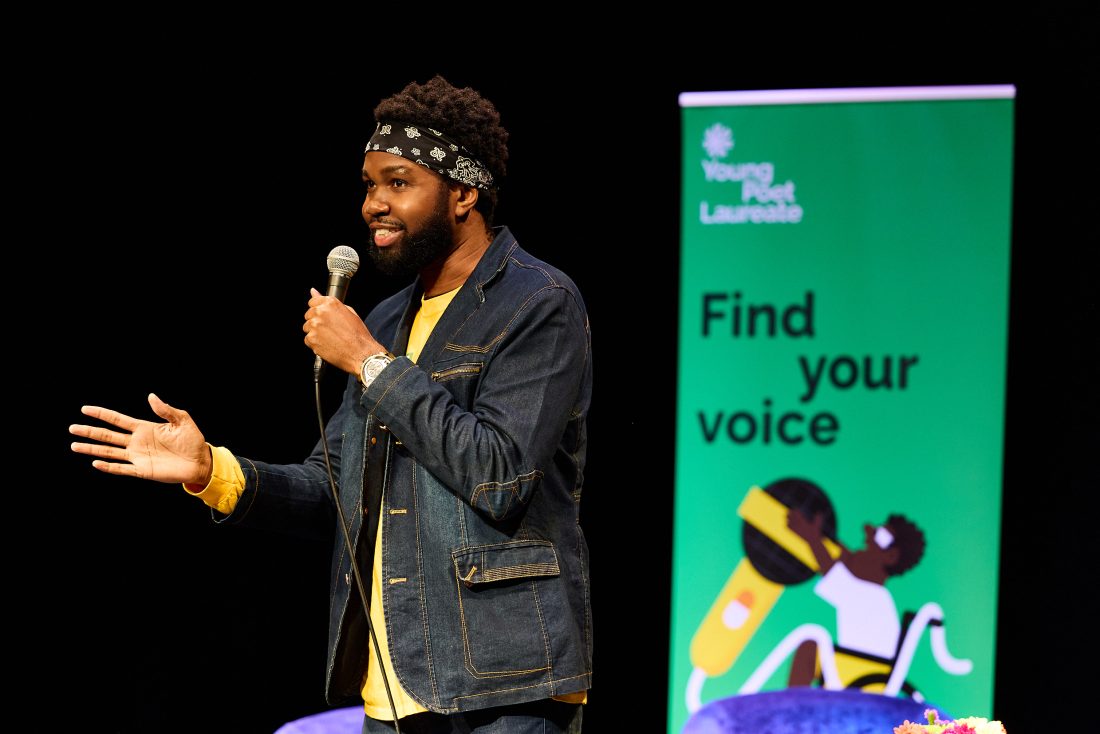 Photo of a young man in a denim jacket, speaking into a microphone. A green banner behind him reads, ‘Find your voice’.