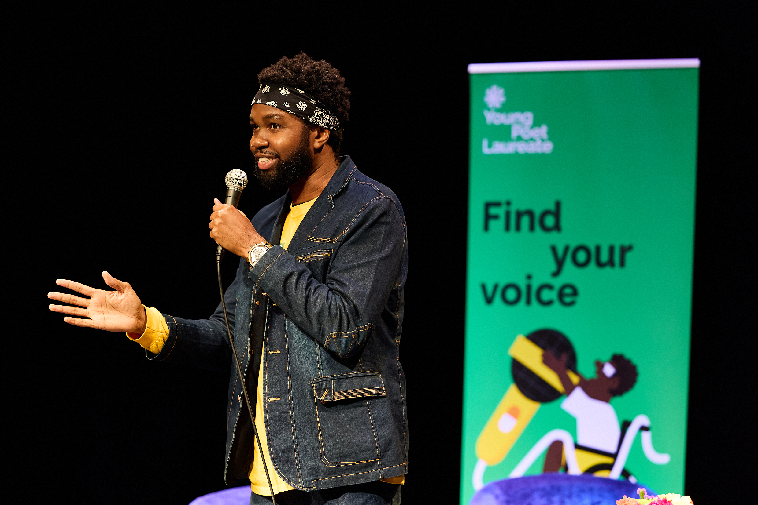 Photo of a young man in a denim jacket, speaking into a microphone. A green banner behind him reads, ‘Find your voice’.