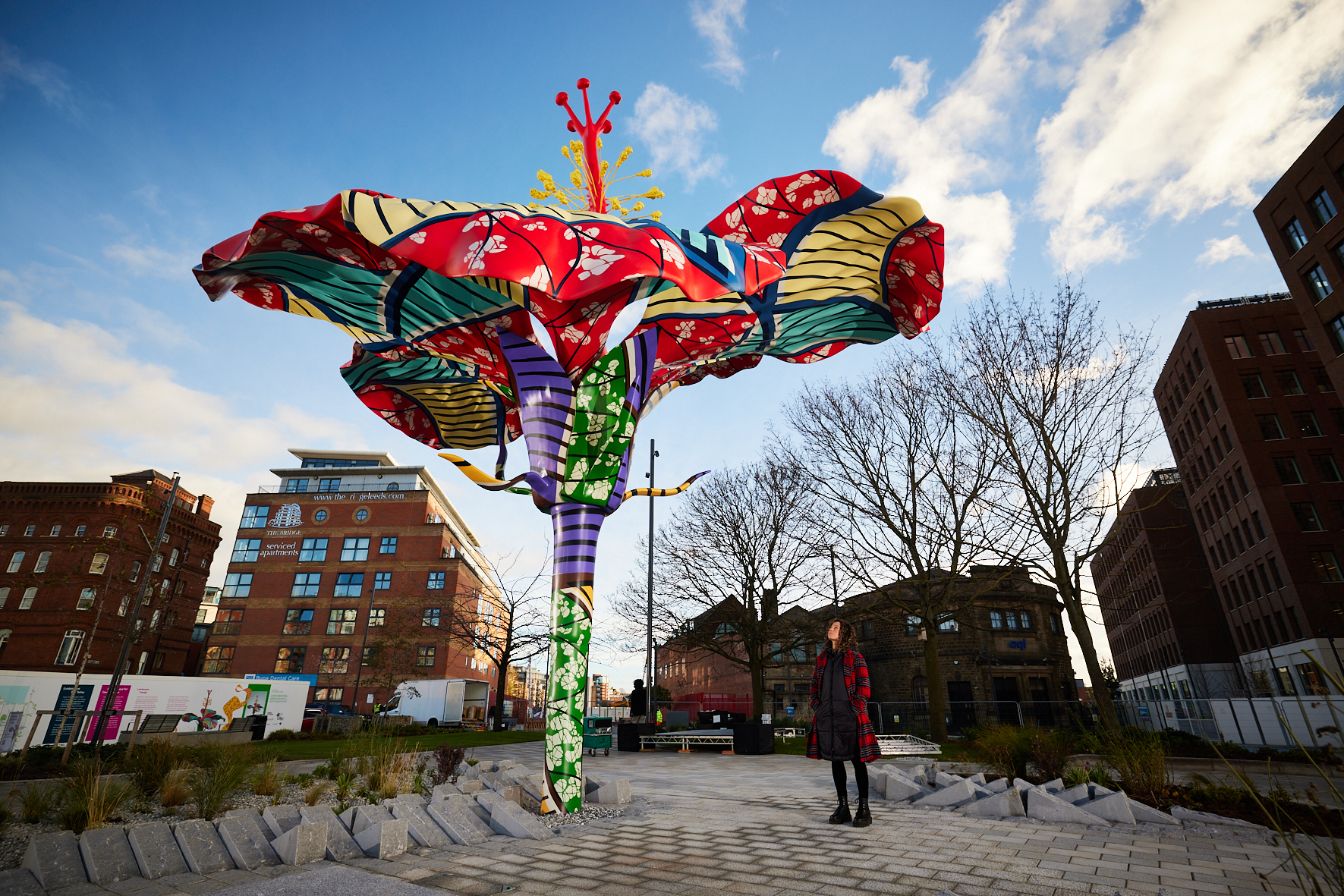 Photo of a woman looking up at an abstract sculpture of a giant and colourful hibiscus flower, with urban buildings in the background.