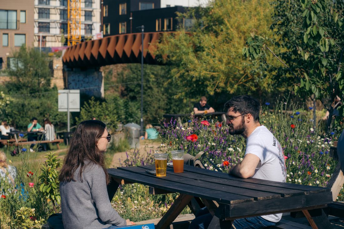Photo of a woman and a man drinking beer on a picnic bench on a sunny day, surrounded by wild flowers, with urban buildings in the background.