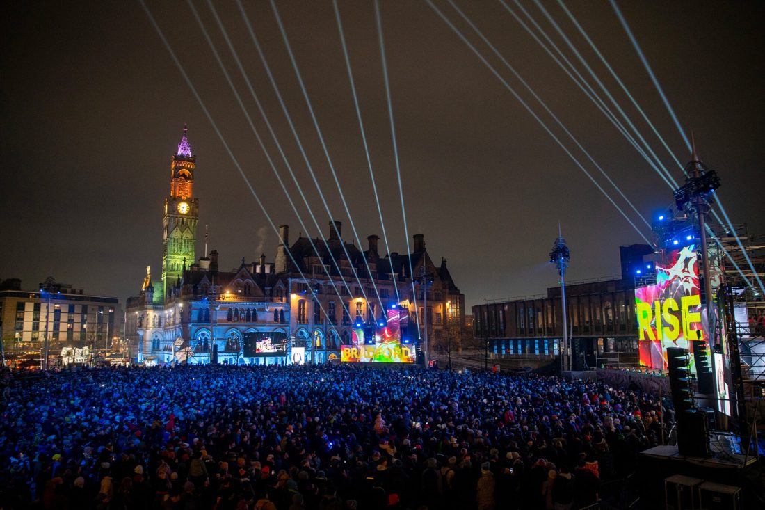 Photo of Bradford city centre at night, packed with crowds. Lazer beams and colourful projections light up the sky and buildings. A huge screen reads, ‘Rise’.