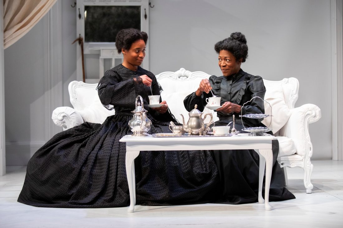 Photo of two Black actors on stage and in character, wearing black, Victorian-style dresses, sitting on a white sofa and stirring teacups.