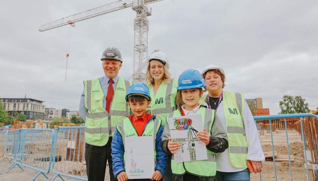 Photo of three adults and two children, all in fluorescent jackets, smiling at the camera. The children are holding drawings of cranes and in the background is a real crane.