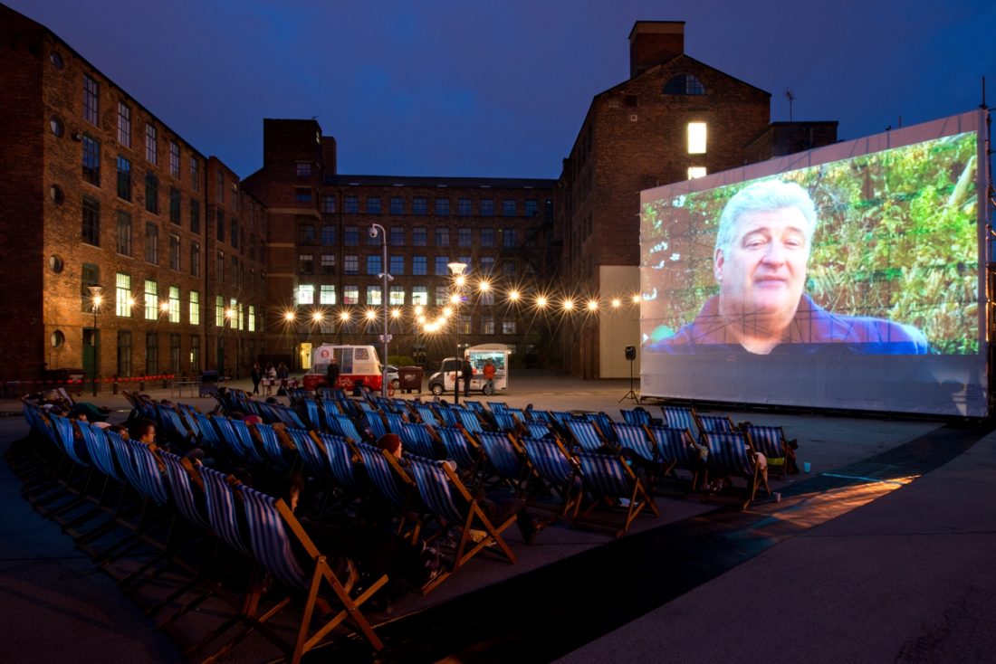 Photo of an outdoor movie screening at dusk in Holbeck Urban Village, with rows of deck chairs facing a screen and converted warehouses in the background.