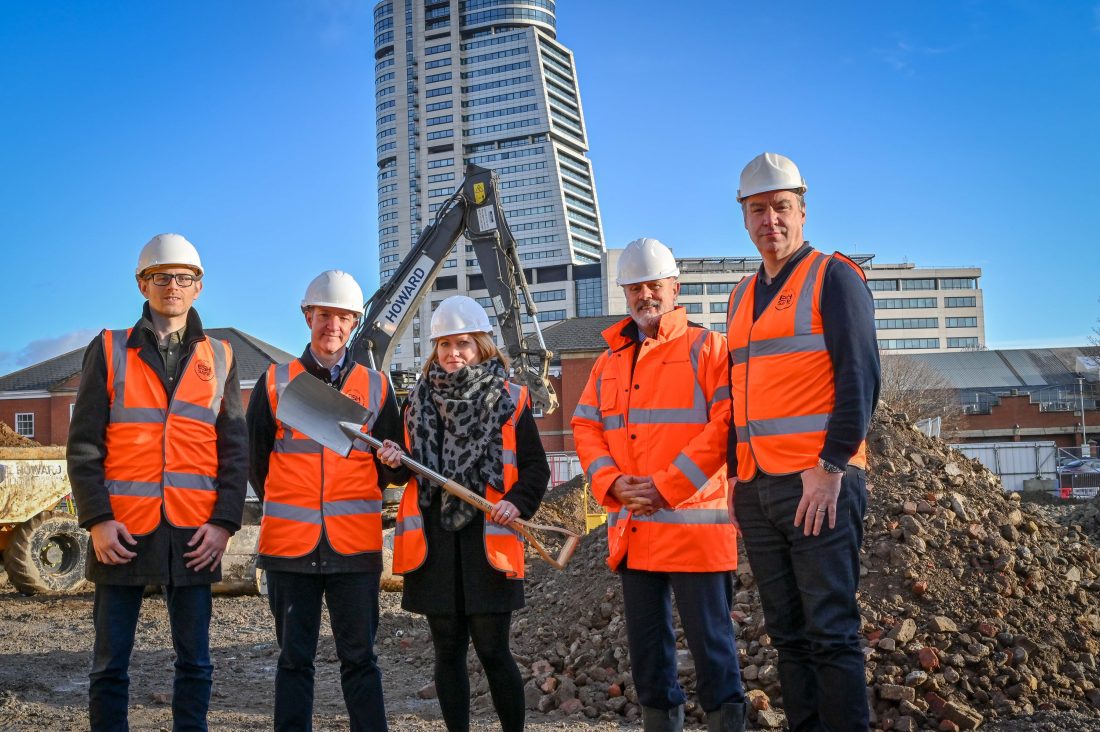 Photo of four people wearing orange fluorescent jackets on a building site. The woman in the middle holds a spade and everyone is smiling at the camera.