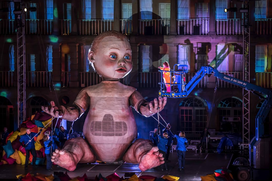 Photo of a giant, 3-D model of a baby sitting in front of one the walls in The Piece Hall at night. People are manoeuvring its hands with poles from the ground, while another person is lit up in a boom lift, facing the baby.