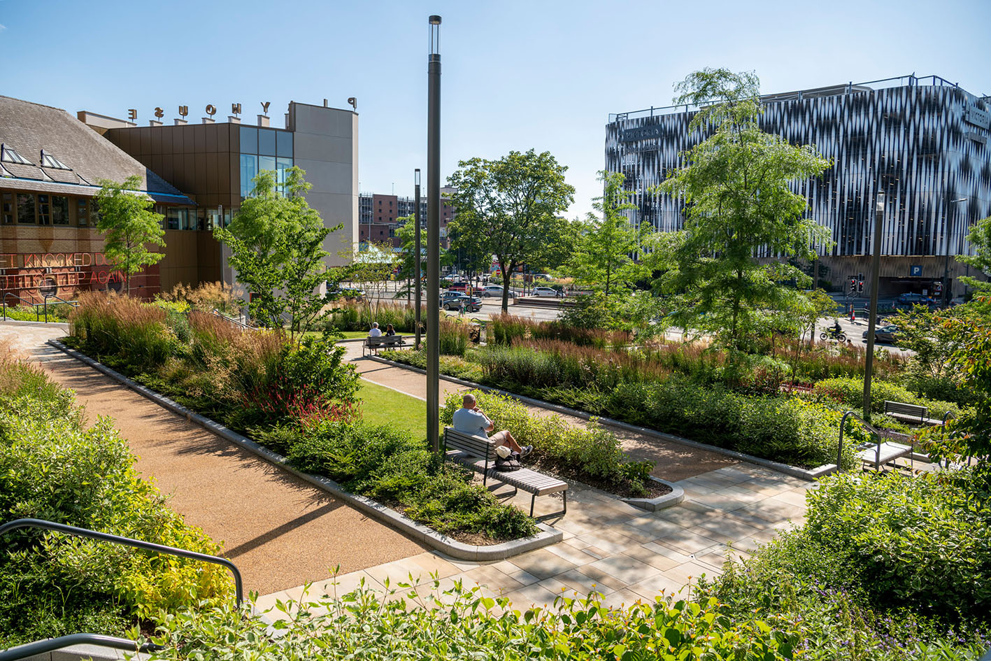 Photo of a communal, urban garden on a sunny day, planted with trees and shrubs. There are people sitting on the benches around the garden.
