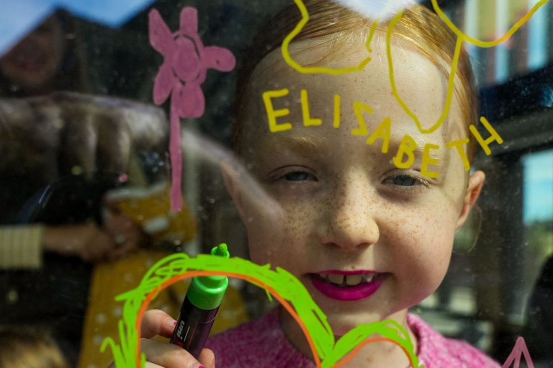 Photo of a little girl in a pink jumper drawing a heart with a face on a window. The photo is taken from outside, looking in.
