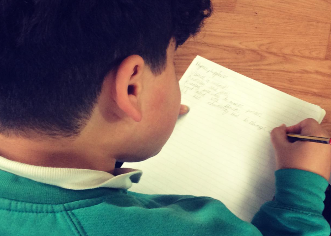 A child sits at a table writing in a lined notebook, seen from behind with their head and shoulders visible.