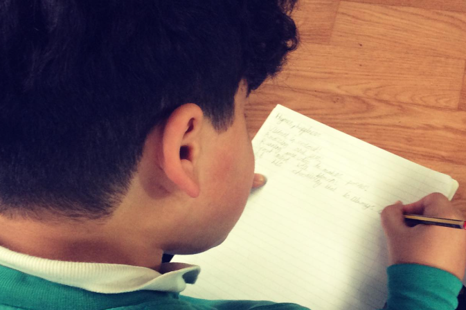 A child sits at a table writing in a lined notebook, seen from behind with their head and shoulders visible.